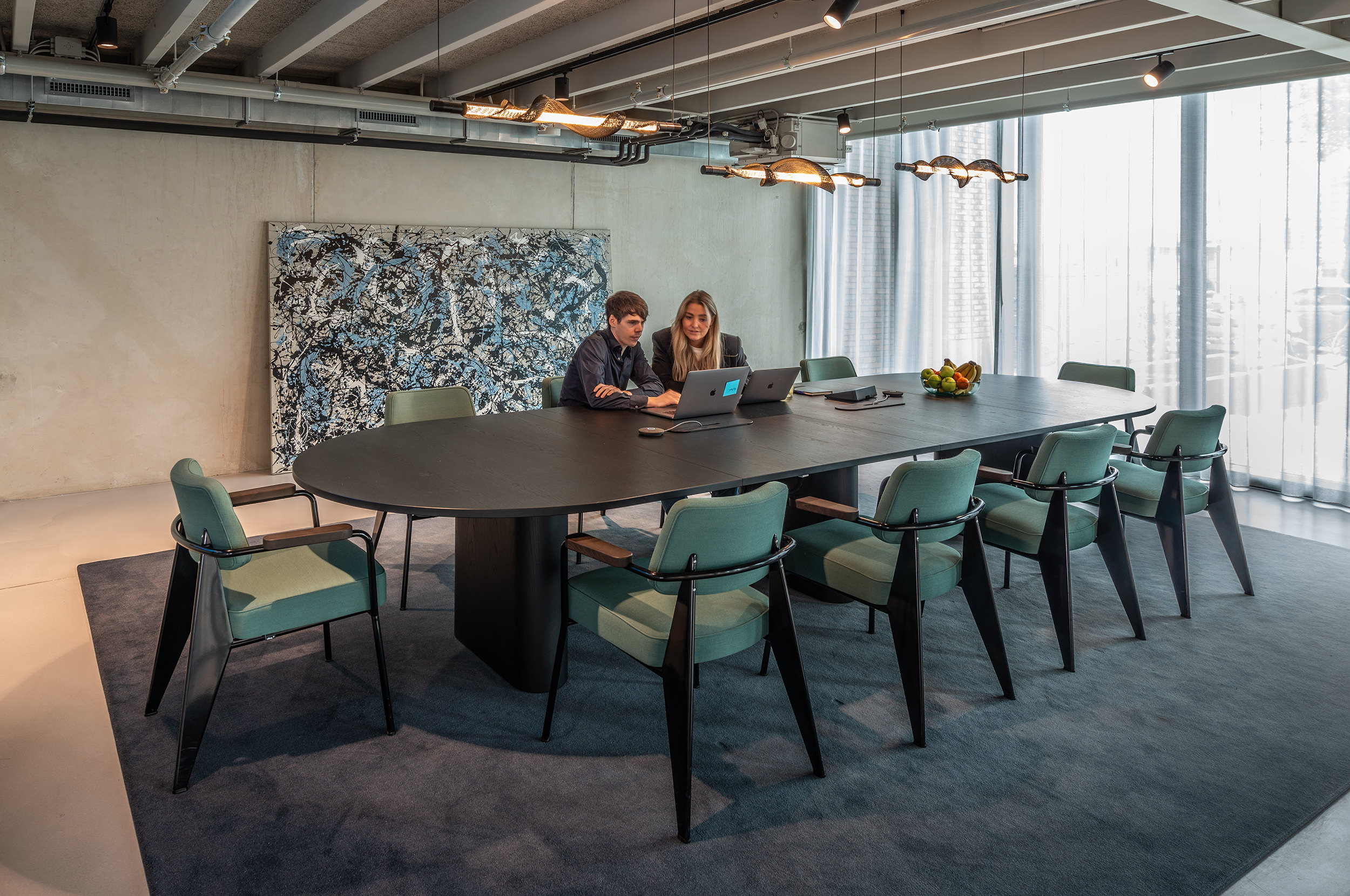 Meetingruimte met langwerpige zwarte ovalen tafel met tien groene stoelen eromheen waar twee mensen aan zitten te werken.