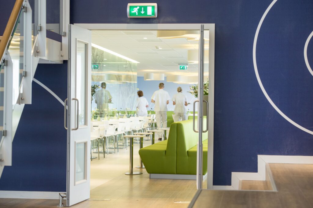 Sfeerafbeelding met doorkijk door openslaande deuren in ruimte met groene banken in het Isala Ziekenhuis. De ruggen van drie doktoren in witte pakken zijn te zien op de afbeelding.