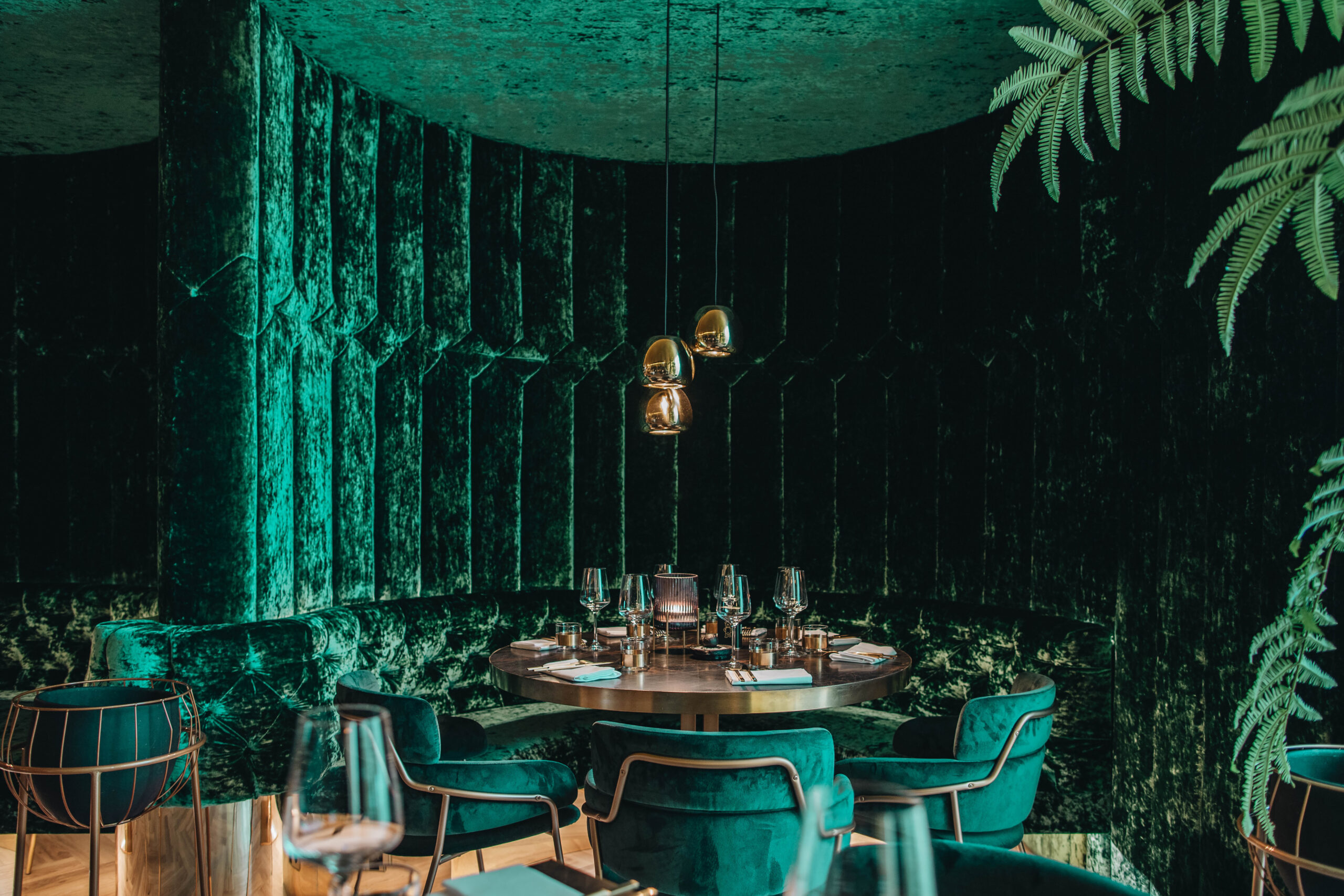 Zithoek in Restaurant Goud met volledig groene, suede, ronde wand en daarin een ronde tafel met dezelfde groene stoelen. Drie hanglampen uit het plafond.