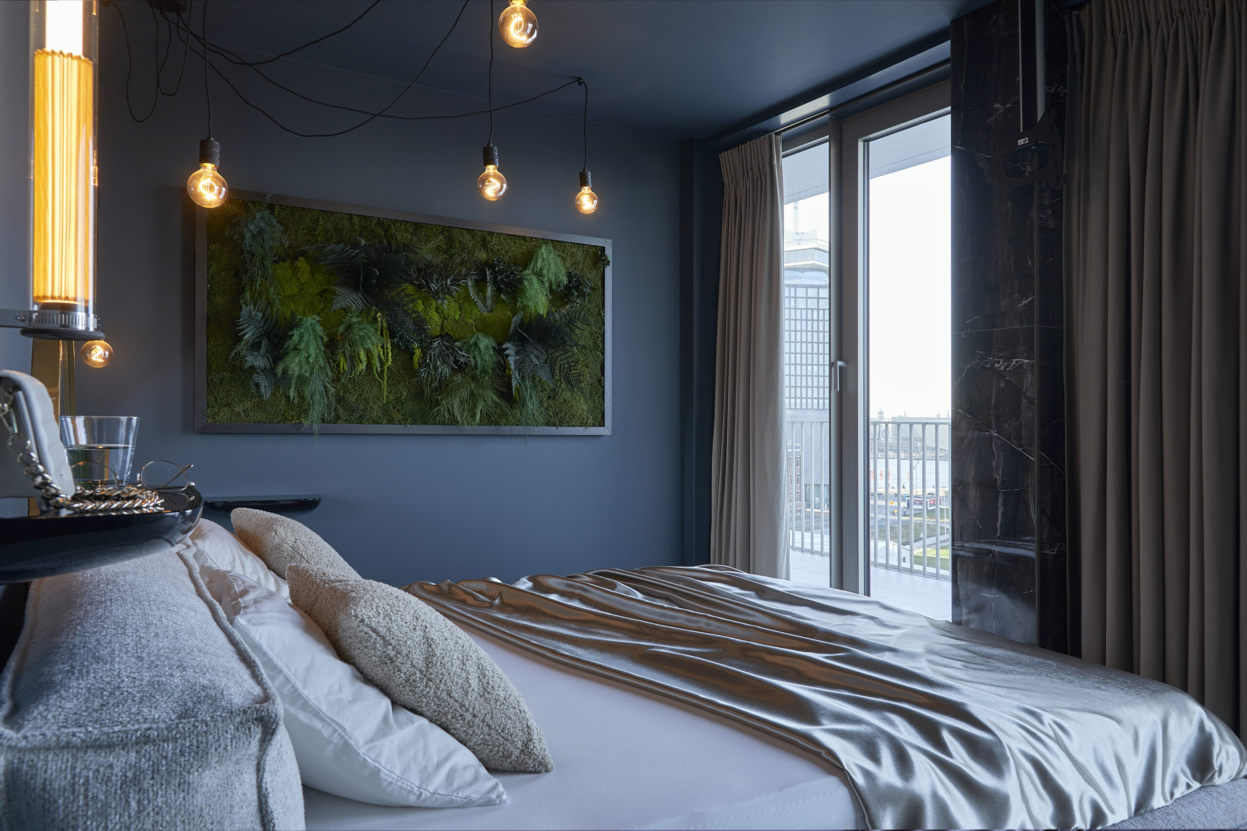 Slaapkamer met ruim bed met satijnen laken, hanglampen aan het plafond en een groen kunststuk aan de muur. Balkon met uitzicht op de stad.