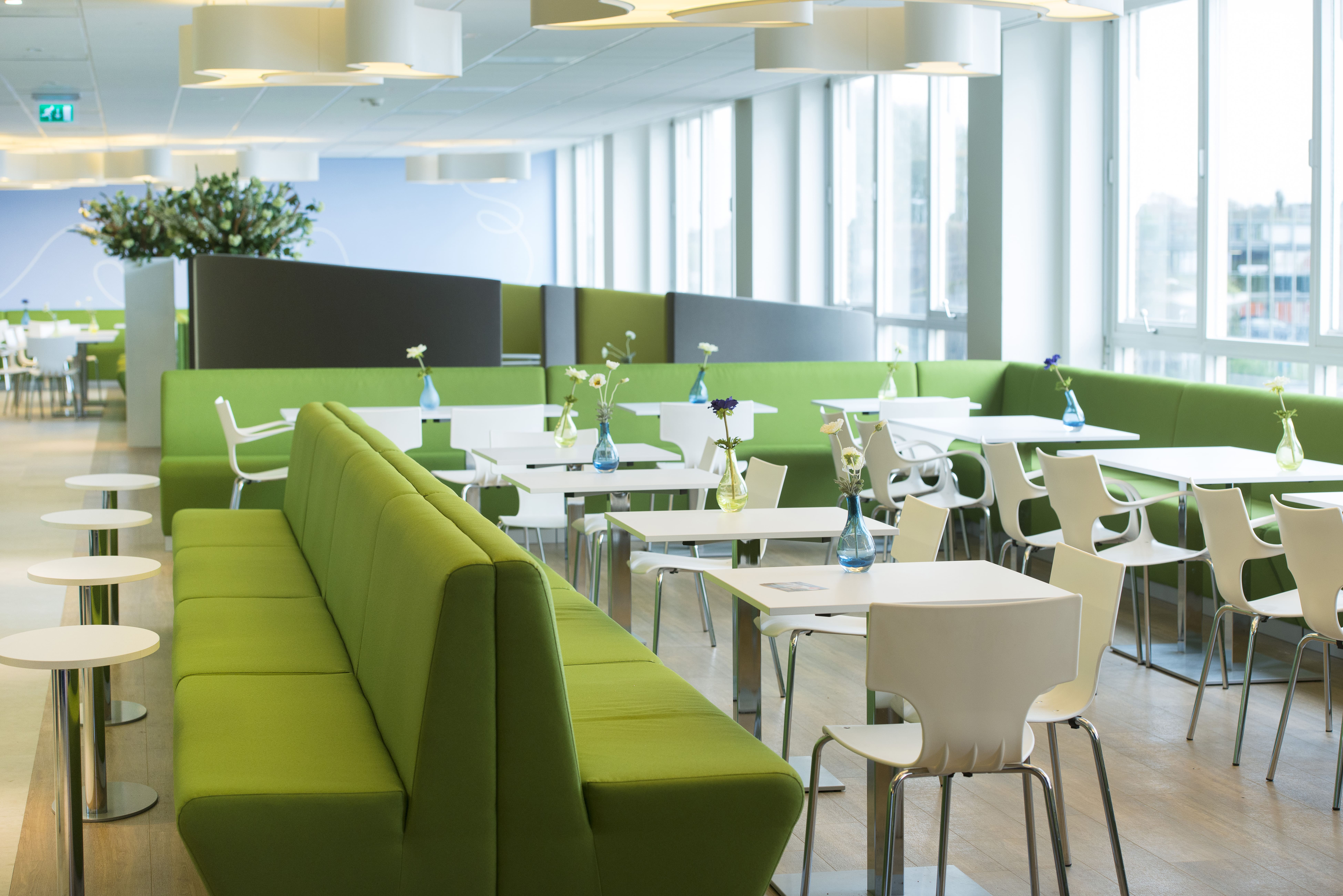 Ruimte in het Isala Ziekenhuis ingericht door Big Brands met lange groene banken met witte tafels en stoelen. Bloemen op de achtergrond en witte lampen aan het plafond.