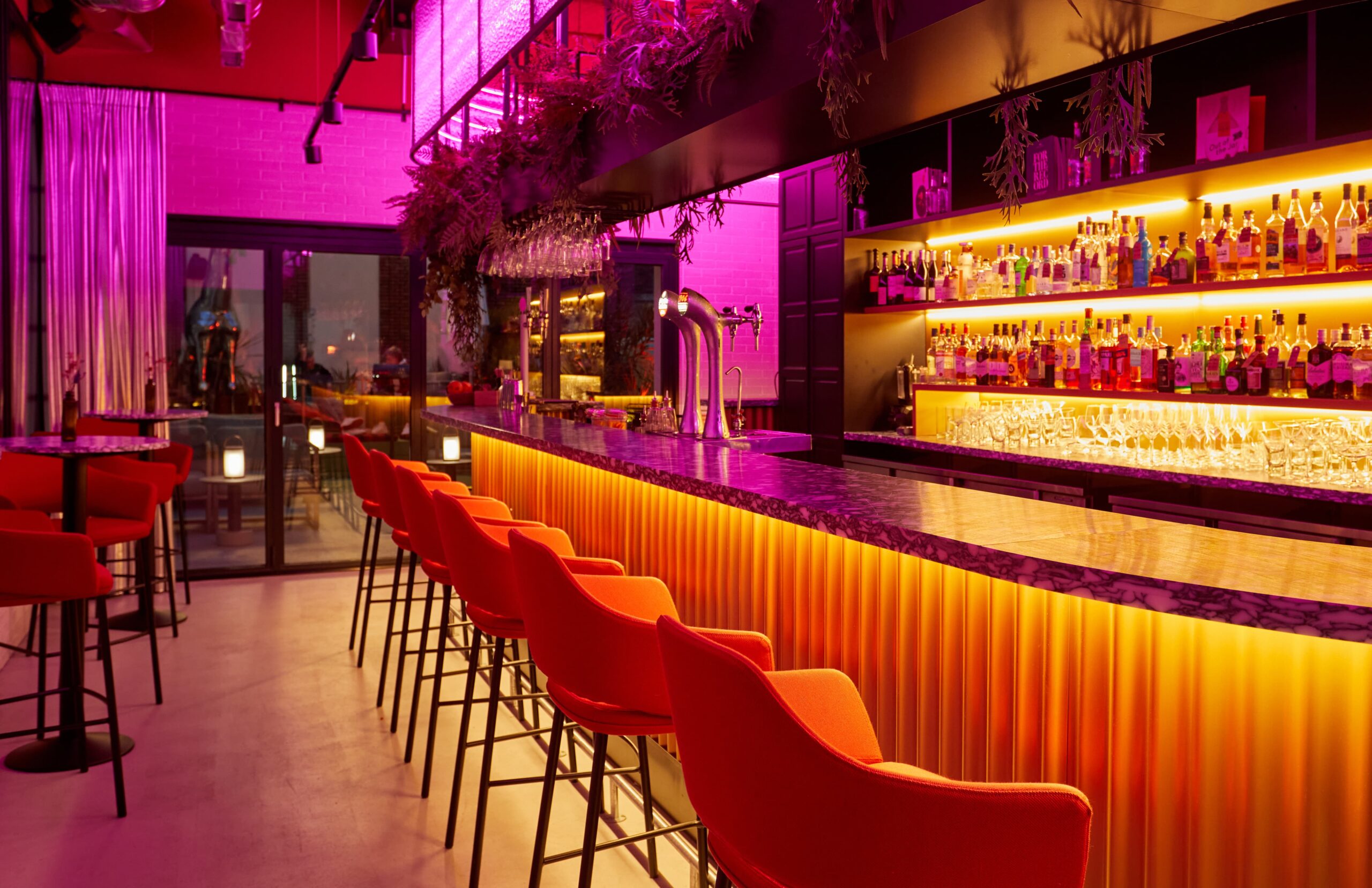 Bar met oranje en roze lichten, hoge barkrukken.