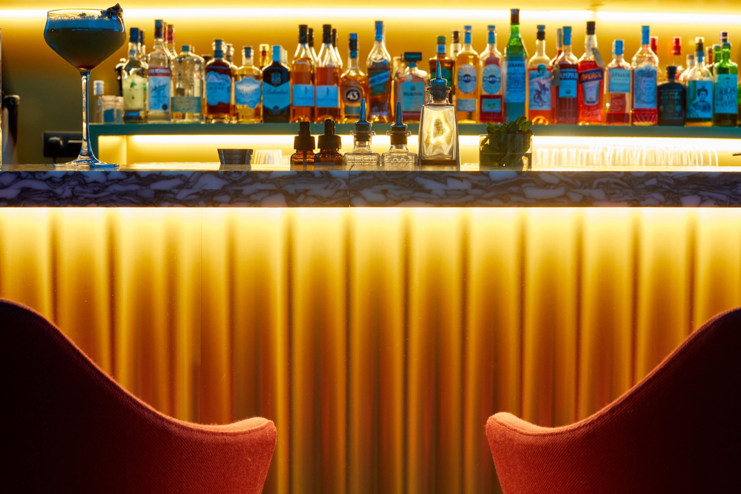 Sfeerafbeelding van de bar met op de voorgrond twee barkrukken en de bar op de achtergrond met veel flessen drank en een cocktail op de bar.