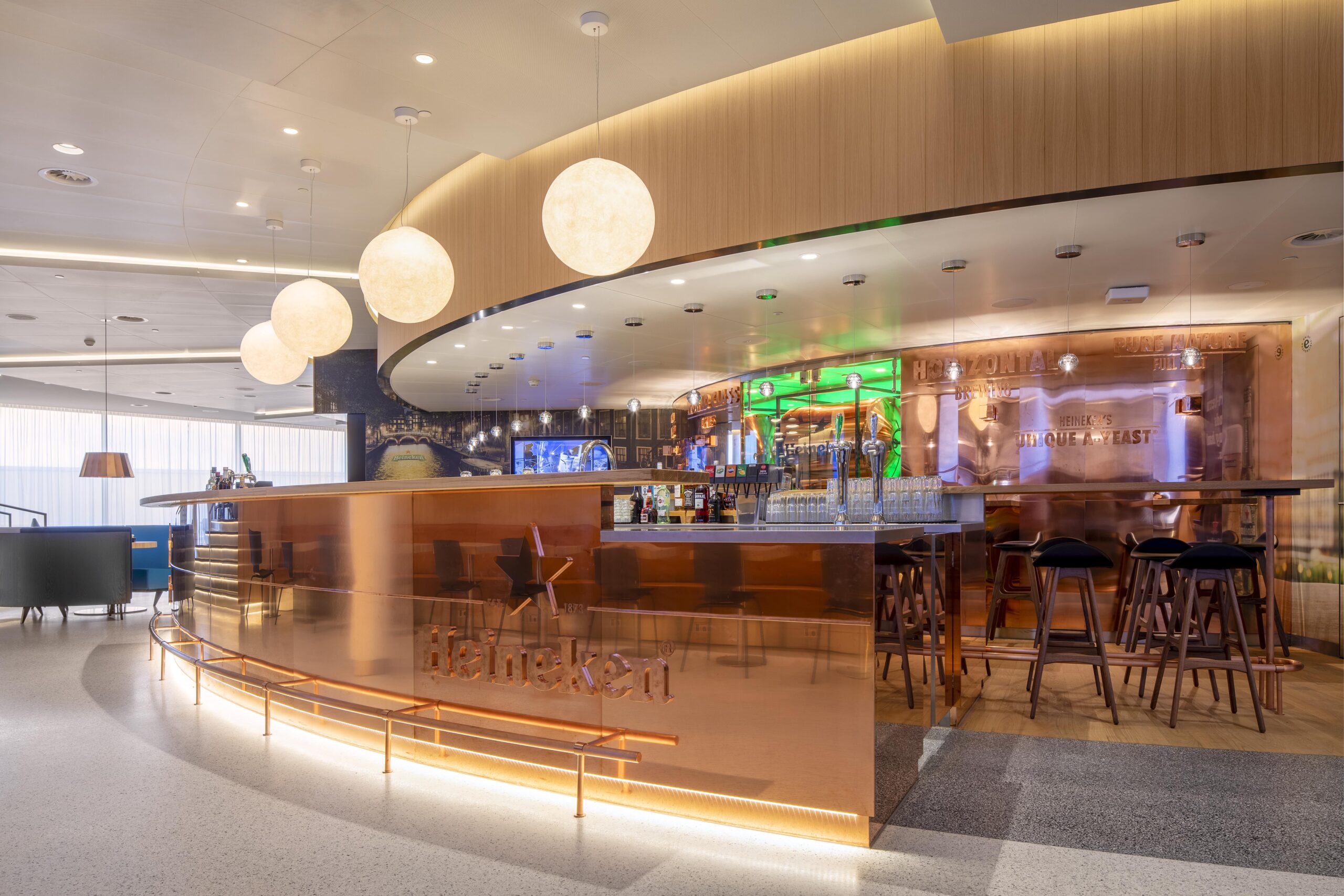 Bar in KLM crown lounge op Schiphol. Met groot Heineken logo en hoge barkrukken. Grote ronde lampen aan het plafond.